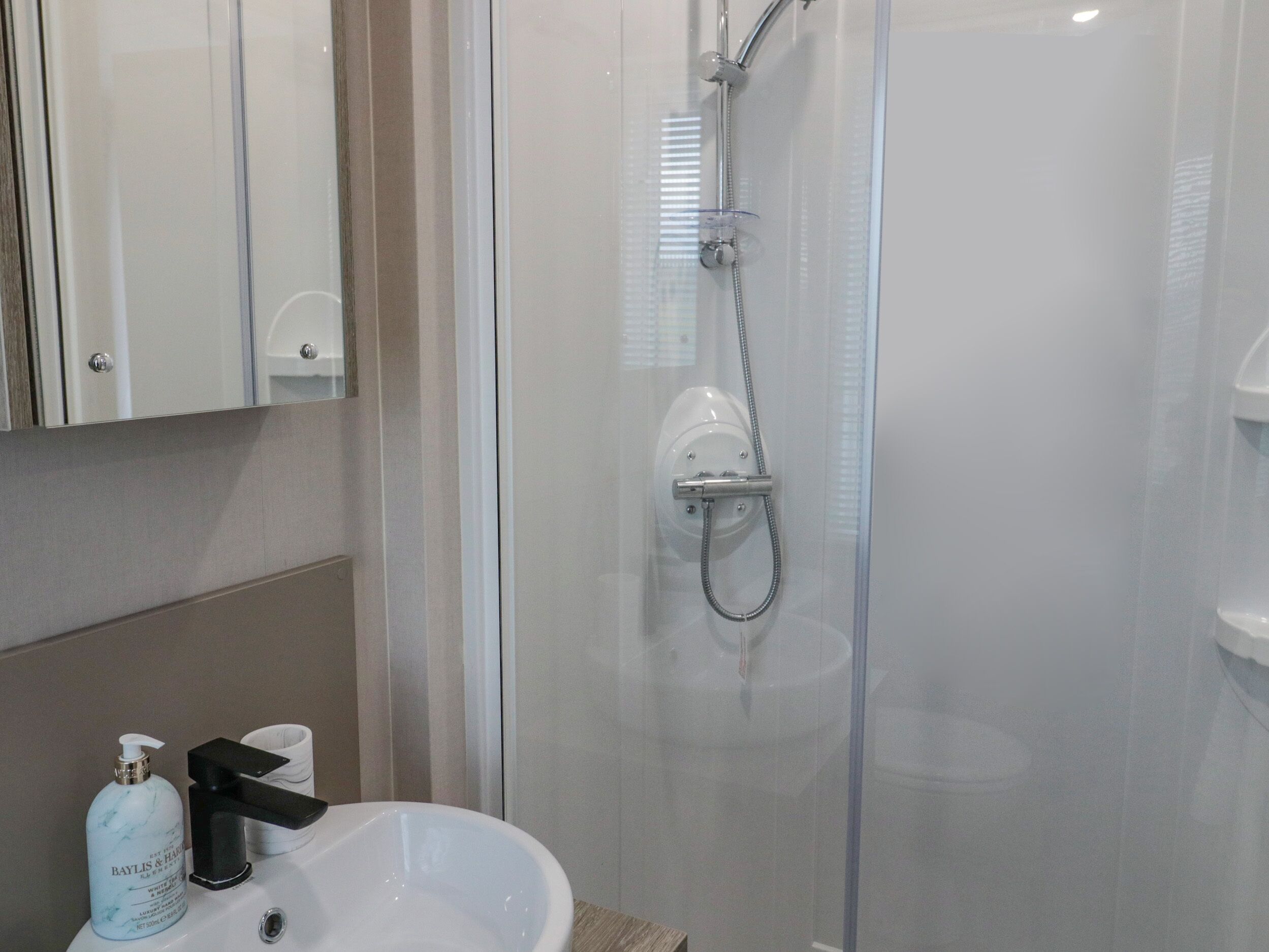 A bathroom with a glass-enclosed shower, a white sink, and a bottle of Baylis & Hensley body wash.