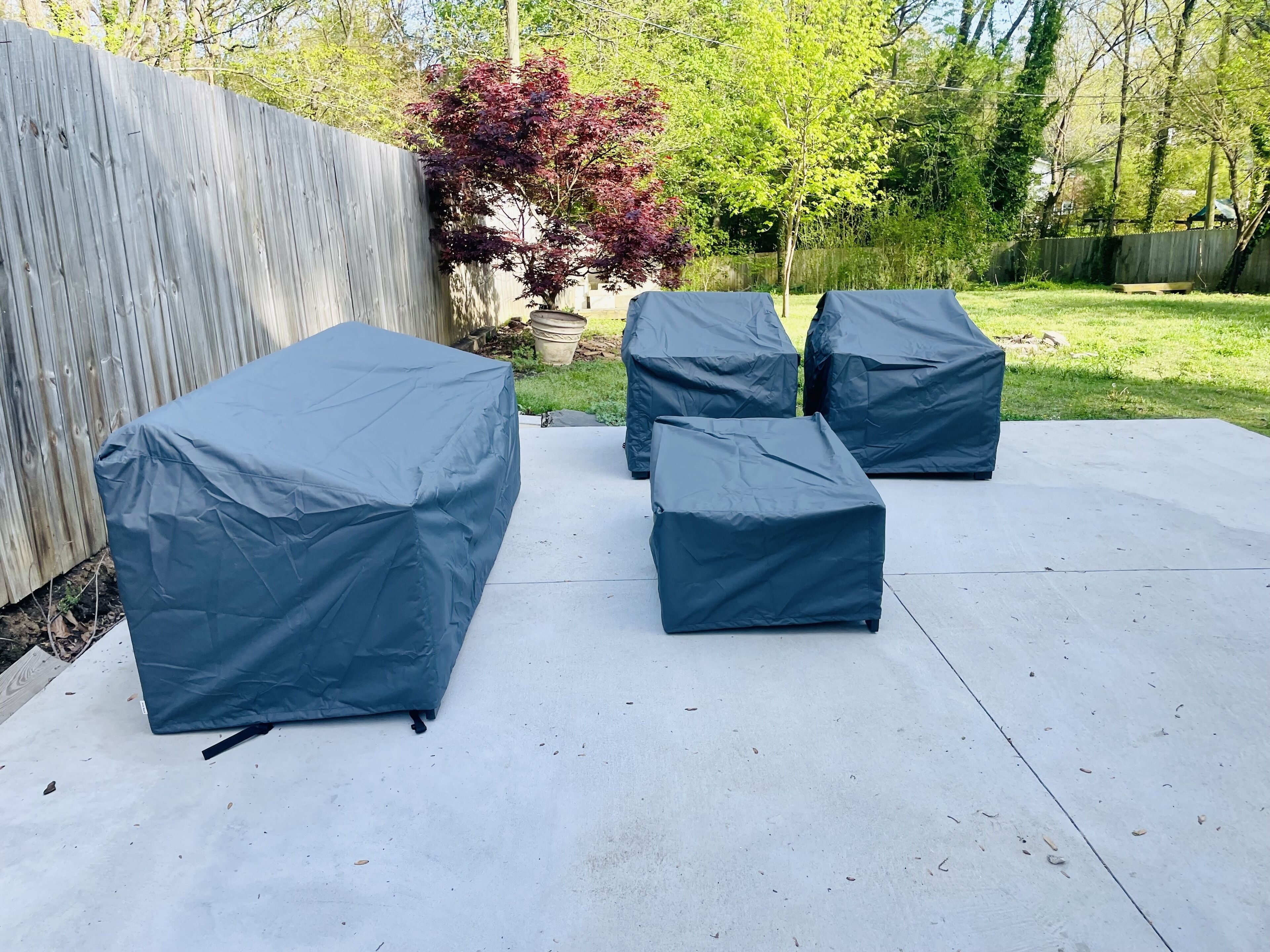 Mobiliario de patio al aire libre cubierto con tela azul, una valla de madera, y un jardín con árboles y un banco al fondo.
