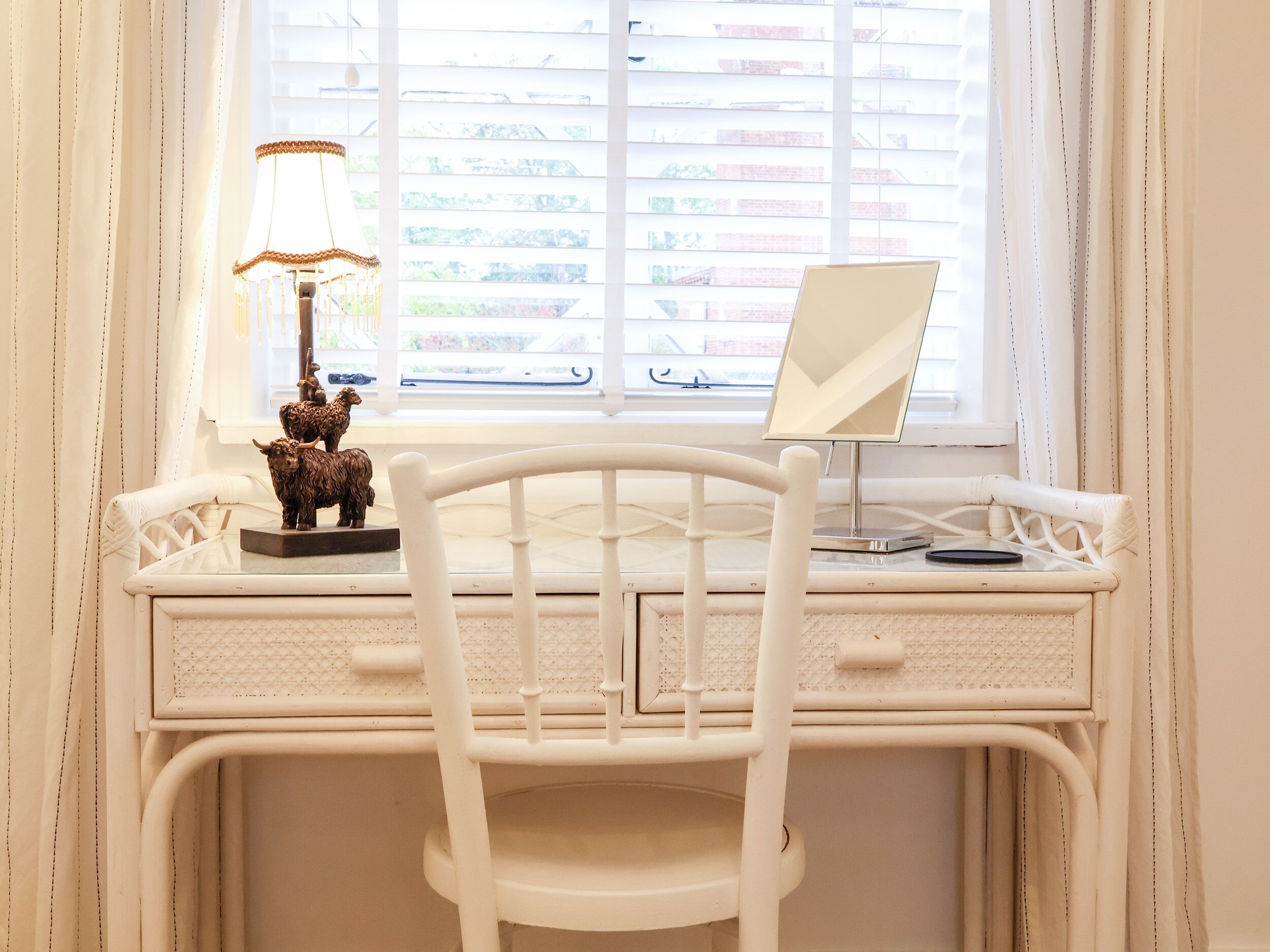 A white desk with a computer and a lamp, a chair, and a window with blinds.