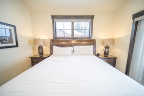 A neatly made bed in a room with a large window, two bedside tables with lamps, and a framed picture on the wall.