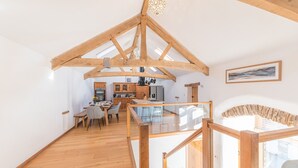 A spacious loft with wooden beams, a kitchen, dining area, and a staircase leading to a balcony. - Swallow Barn (Cockermouth)