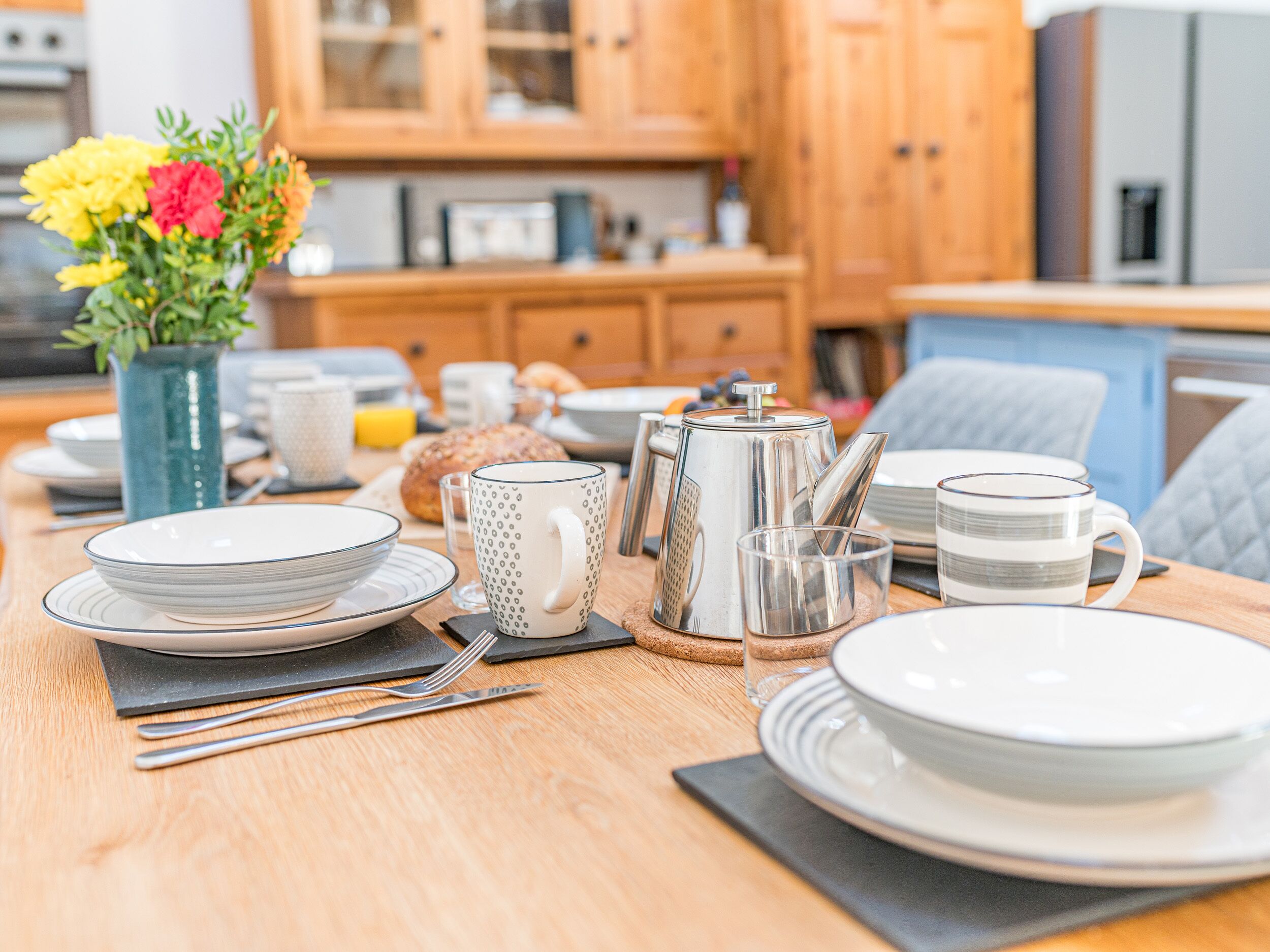 Una mesa de desayuno dispuesta con platos, tazas y una tetera, con un jarrón de flores al fondo.