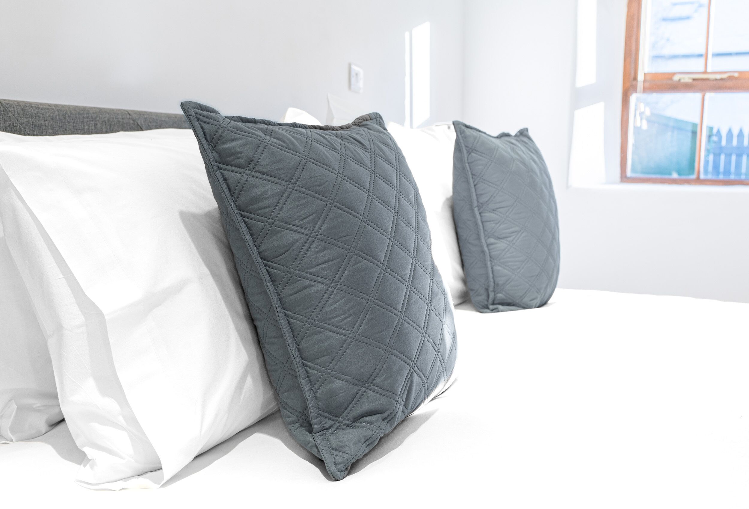 A bed with white and gray pillows, a window with a view of a fence, and a white wall.