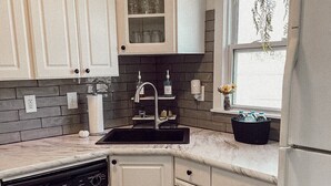 Una cocina con armarios blancos, un lavavajillas negro, un lavamanos con grifo y una ventana con una planta.
