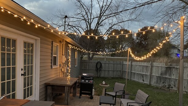 Un patio trasero con parrilla, mobiliario exterior e iluminación con luces colgantes.