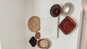 Un banco de madera pequeño con un cojín tejido, una pared con cestas tejidas decorativas y una obra de arte enmarcada.