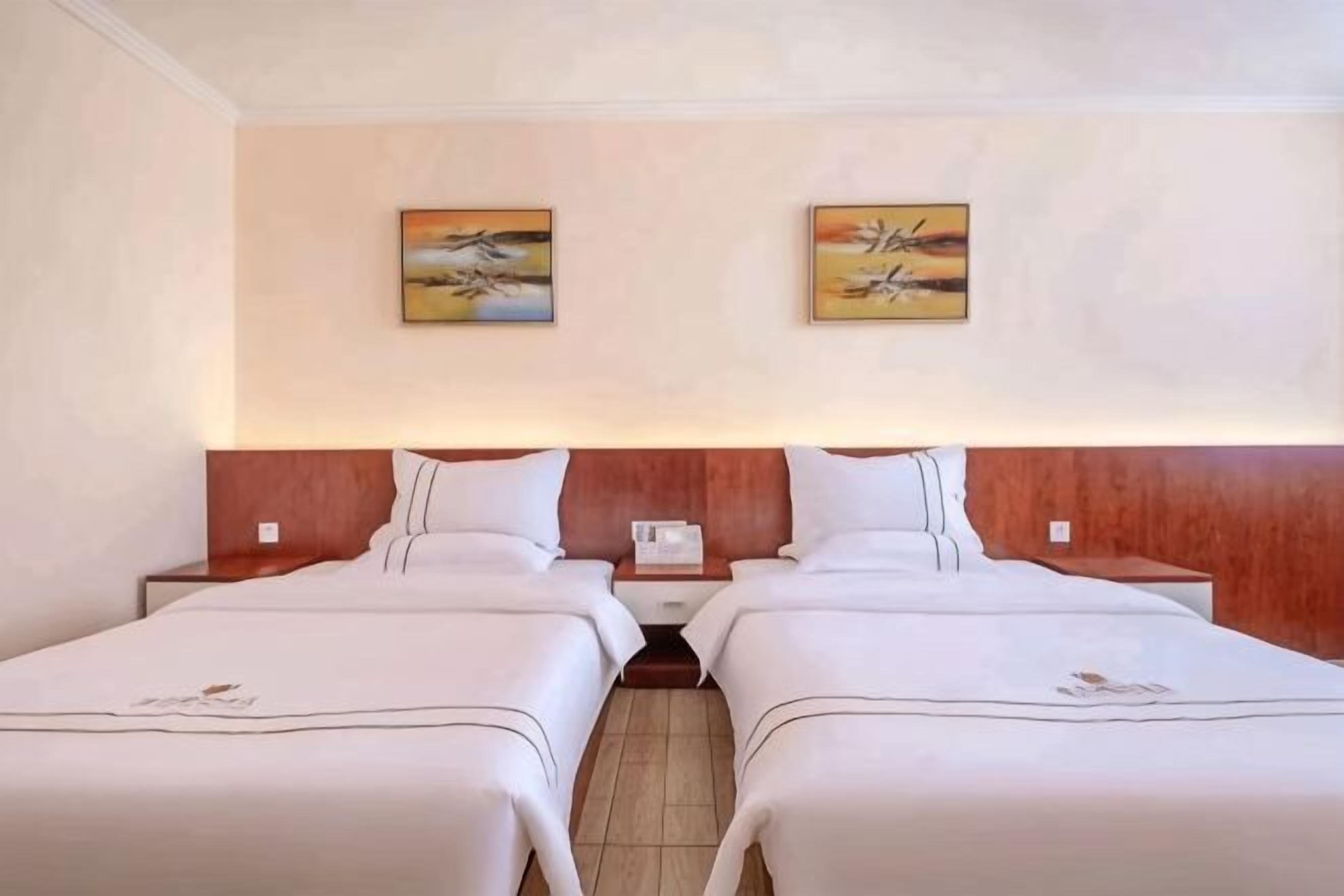 A hotel room with two beds, wooden headboards, artwork on the walls, and a nightstand with a phone.