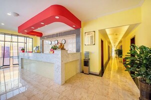 Una zona de recepción de hotel con mostrador de mármol, pared decorada en rojo y un largo pasillo con plantas en macetas.