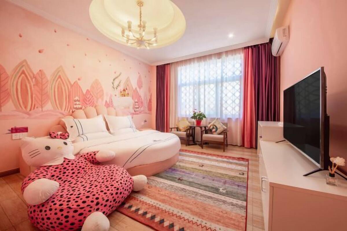 A bedroom with a bed, a TV, a window with curtains, a small table with a plant, and a decorative wall.