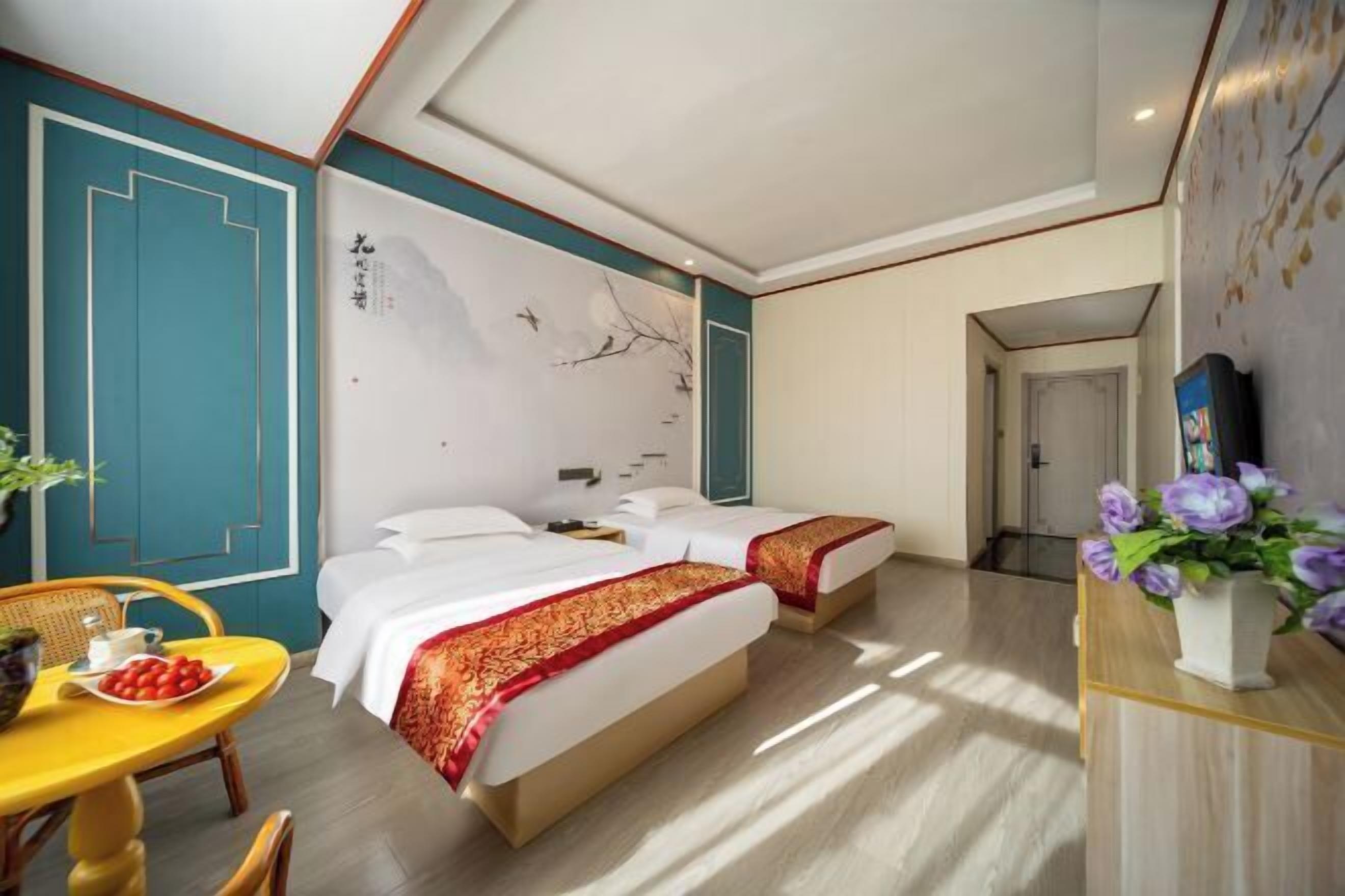Ett hotellrum med två sängar, ett matbord med stolar, en vas med blommor och en vägg med en dekorativ panel.