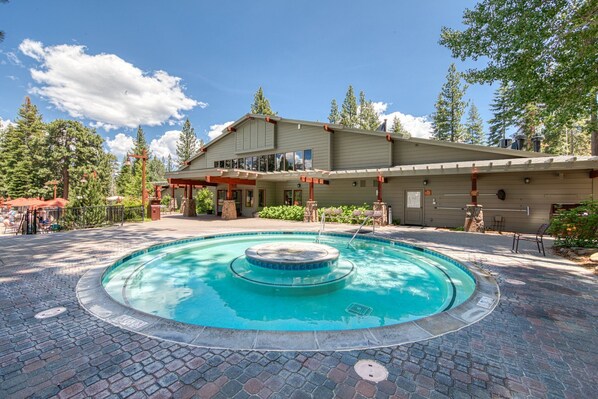 A pool area with a hot tub, surrounded by a brick patio, and a building with a covered deck and outdoor seating.