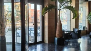 Eine Lobby mit Glastür, einer Palme und einer Sitzecke mit Stühlen und Tischen.
