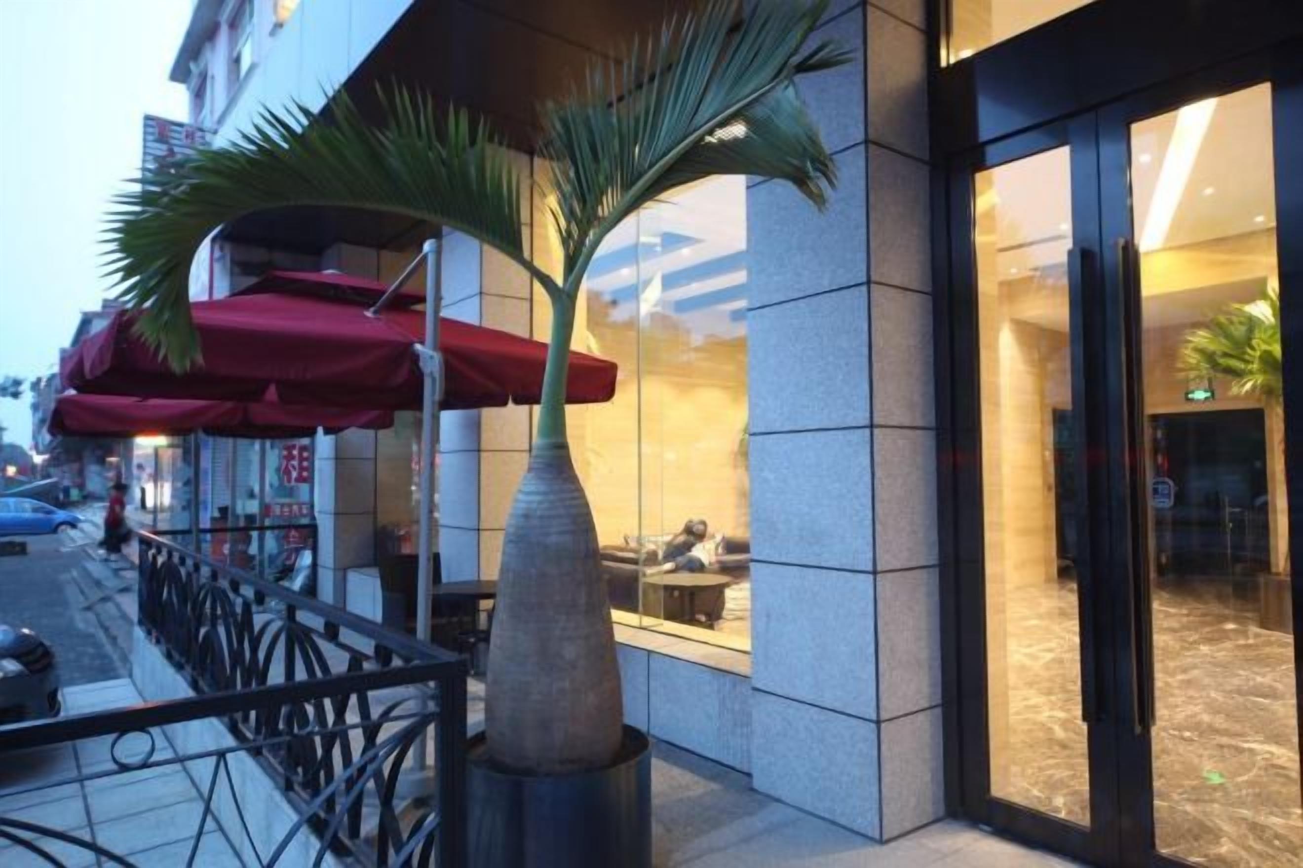 Un edificio con entrada de vidrio, mesas al aire libre con sombrillas rojas y una palmera.