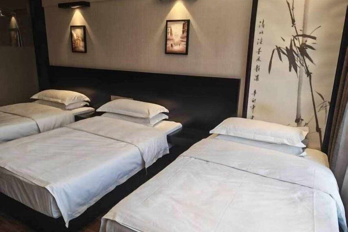 Habitación de hotel con cuatro camas dispuestas en fila, cabecera oscura, obra de arte colgada en la pared y una planta de bambú grande.