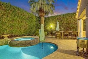 Una piscina con borde de piedra, rodeada por un seto, y una mesa con sillas y sombrillas.