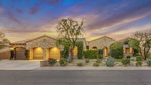 A residential property with a stone facade, multiple garage doors, and a landscaped garden with various plants and a large tree. - Palm Oasis: Pool, Spa, Putting Green, BBQ! (Indio)