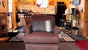 Un salon chaleureux avec un canapé à motifs, un coin repas avec une table et des chaises, et une cuisine équipée d’un réfrigérateur et d’un four à micro-ondes.
