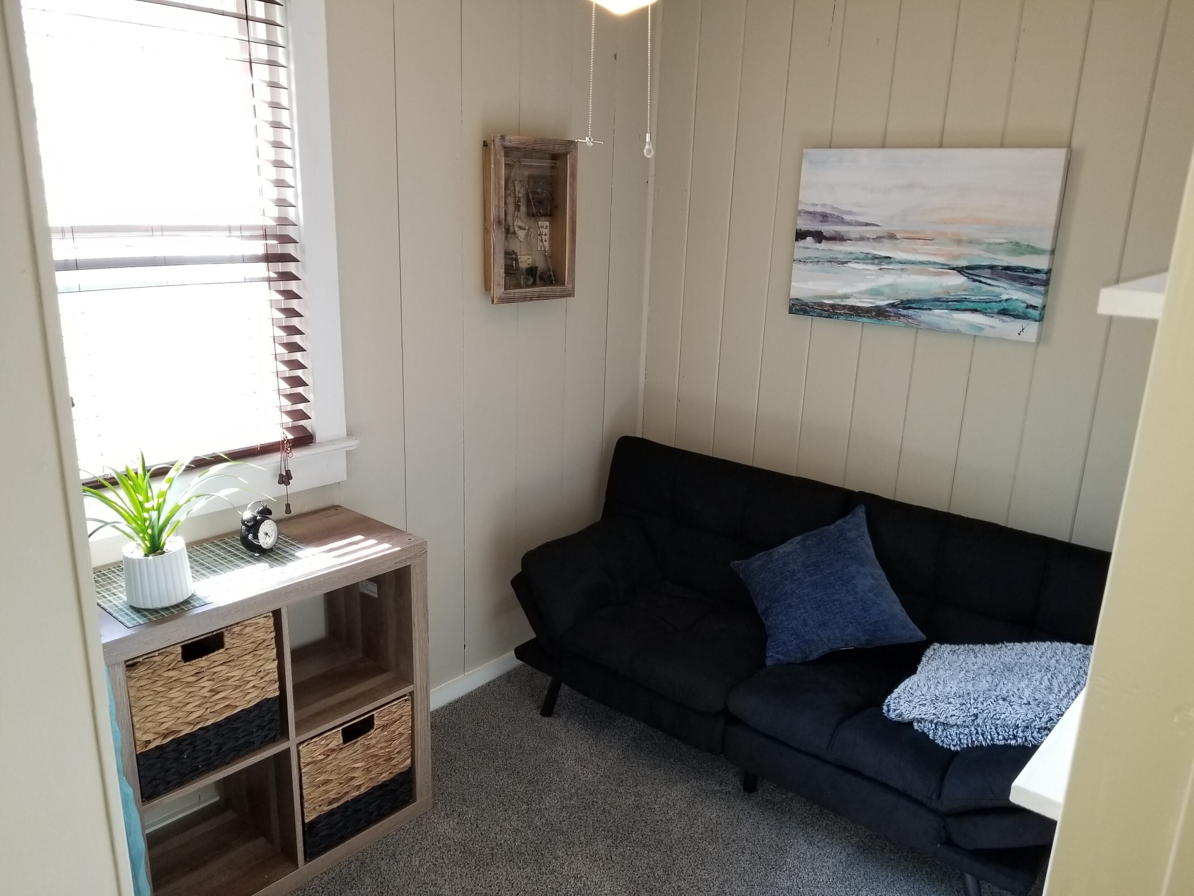 Una habitación acogedora con un sofá negro, una mesita con una planta, un reloj y un cuadro en la pared.