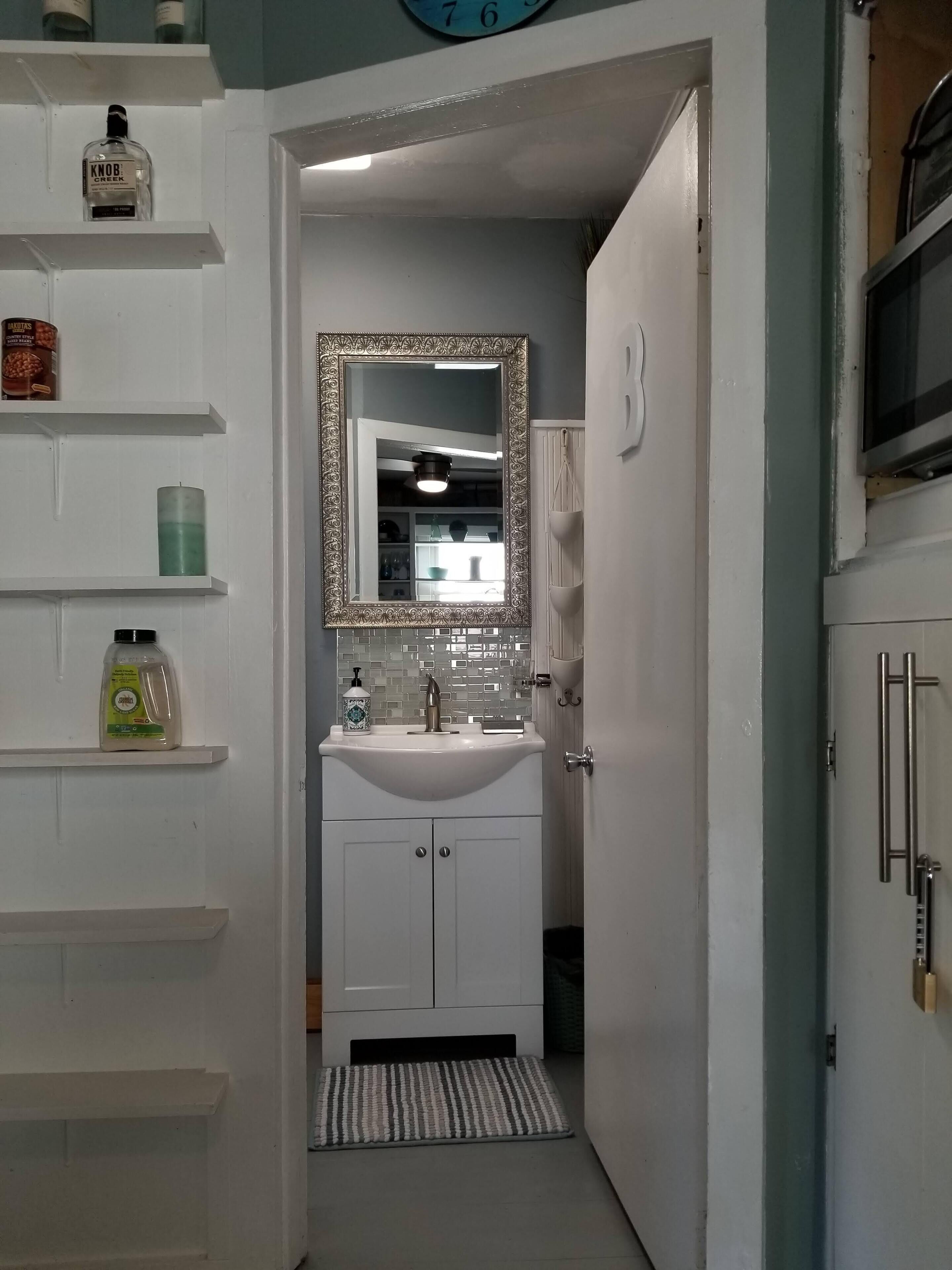Un baño estrecho con un lavamanos blanco, un espejo con un marco ornamentado y estantes con artículos de aseo.