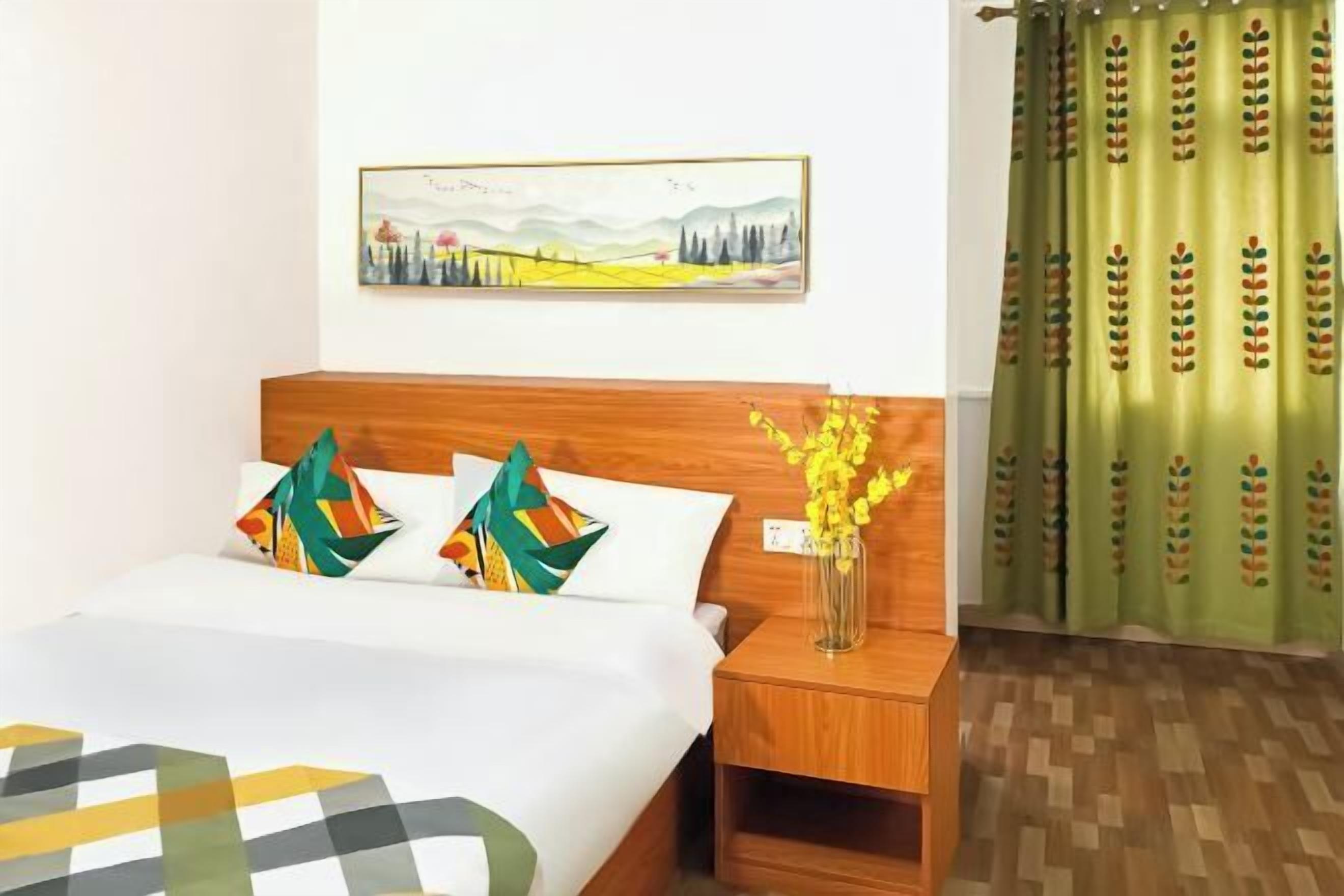 Une chambre d’hôtel avec une tête de lit en bois, un lit recouvert d’une couvre-lit à motifs géométriques, deux coussins colorés, un vase avec des fleurs jaunes, un tableau représentant un paysage encadré accroché au mur et un rideau vert orné de motifs de feuilles.