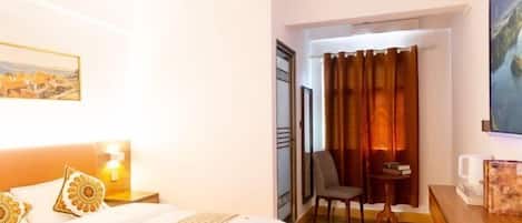 A hotel room with a bed, a chair, a small table, a painting, and a curtain.
