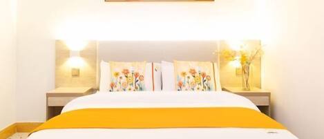 Una habitación de hotel con una cama, dos mesitas de noche, una lámpara de noche, una obra de arte en la pared y un jarrón con flores.