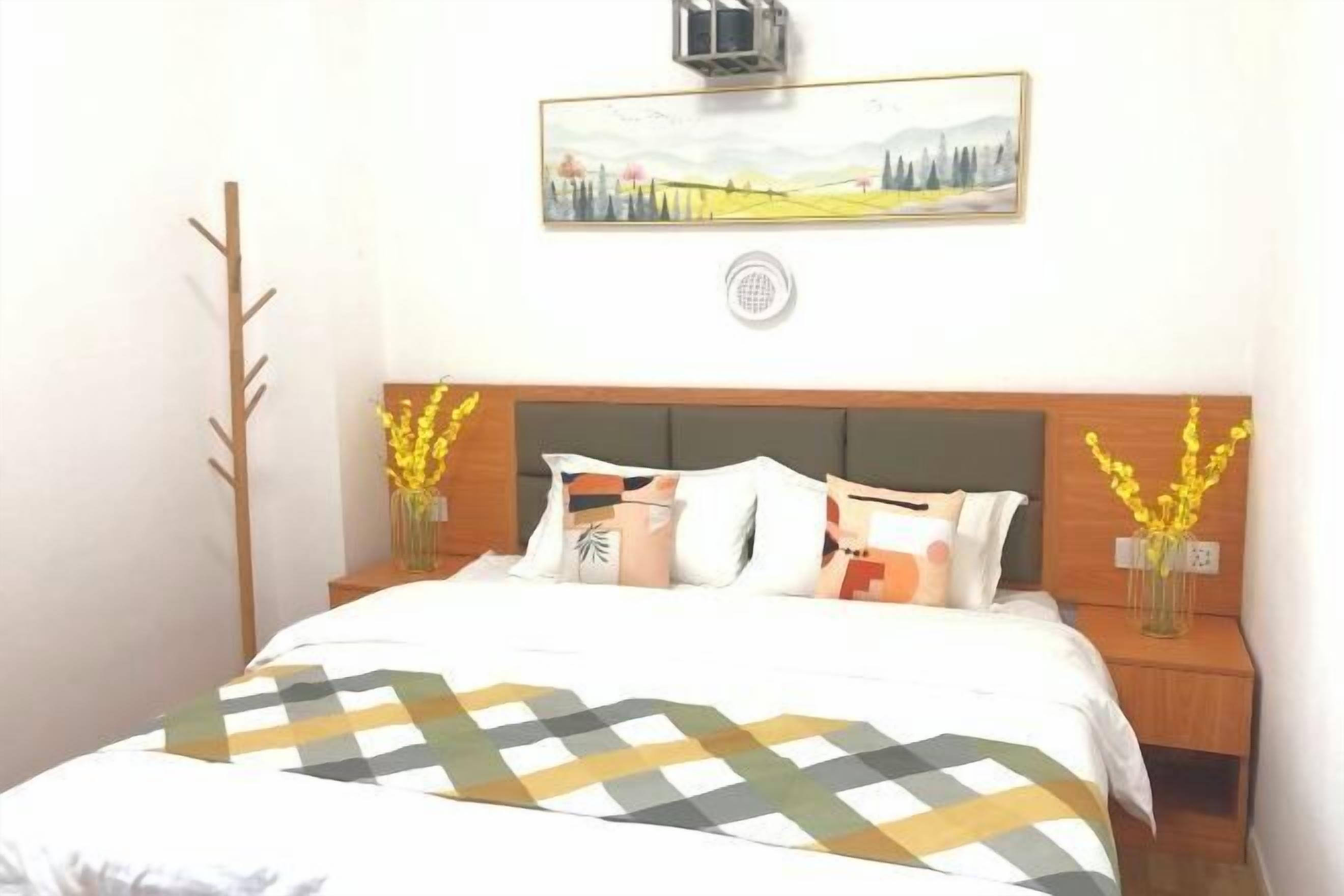 Un lit bien fait, recouvert d’une couvre-lit à motifs, deux tables de chevet ornées de vases remplis de fleurs jaunes, un cadre photo fixé au mur et un petit purificateur d’air également fixé au mur.