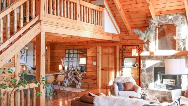 A spacious log cabin interior with a high ceiling, a central fireplace, and a cozy seating area. - Luxury, Curated Lakefront Cabin with New Hot Tub. Near Breweries and Vineyards! (Faber)