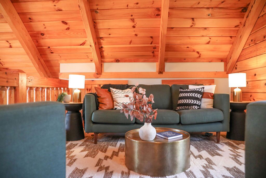 A cozy living room with a wooden ceiling, a green sofa, patterned cushions, a vase with flowers, and two lamps on side tables.