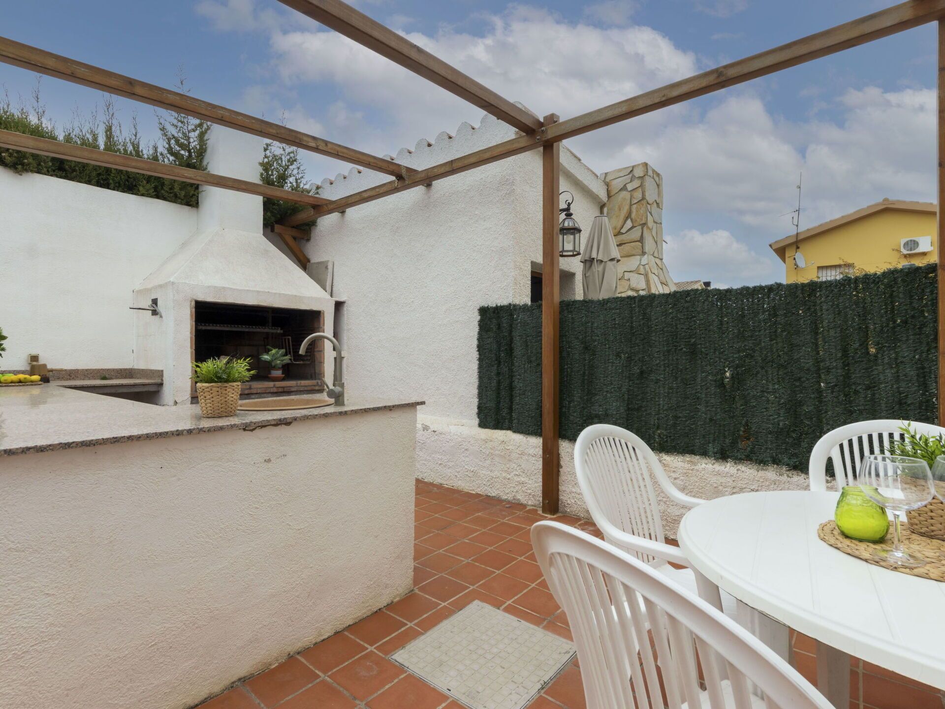 A patio with a built-in grill, white chairs, and a table with a glass and a green vase.