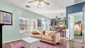 A living room with a beige sofa, a colorful rug, and a white ottoman. There are multiple windows with blinds, a ceiling fan, and a kitchen area visible through an open door. - Stylish Ranch House | Renovated Interior + Backyard Pool Oasis (Dripping Springs)