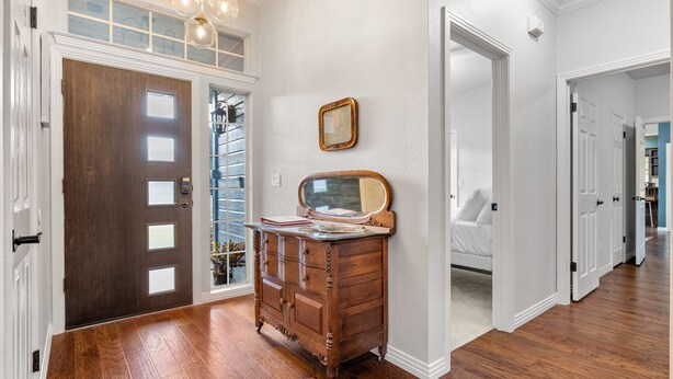 A hallway with a wooden floor, a dark wooden door, a dresser with drawers, a mirror, and a light fixture. - Stylish Ranch House | Renovated Interior + Backyard Pool Oasis (Dripping Springs)