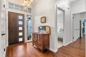 A hallway with a wooden floor, a dark wooden door, a dresser with drawers, a mirror, and a light fixture. - Stylish Ranch House | Renovated Interior + Backyard Pool Oasis (Dripping Springs)