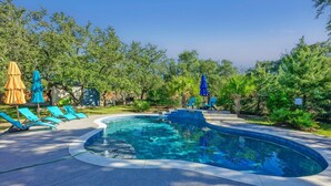 A swimming pool with a curved edge, surrounded by blue lounge chairs and umbrellas. - Stylish Ranch House | Renovated Interior + Backyard Pool Oasis (Dripping Springs)
