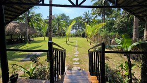 Um deck de madeira leva a uma construção com telhado de palha, com vista para um jardim tropical.