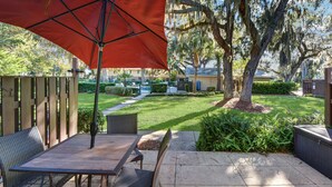 Un espace extérieur aménagé en terrasse, avec une table et des chaises, un parasol rouge, et une pelouse bien entretenue agrémentée d’arbres et d’une clôture.
