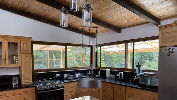 A kitchen with wooden cabinets, a black countertop, a stove, and a sink. There are windows with a view of greenery outside. - Comfortable Cottage for Groups on 2 acre Homestead (San Jose Chacaya)