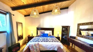 A bedroom with a wooden ceiling, a large bed with a patterned bedspread, two bedside lamps, a bench with a pillow, a mirror, and a bookshelf. - Comfortable Cottage for Groups on 2 acre Homestead (San Jose Chacaya)
