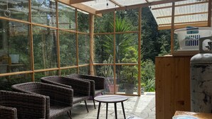 A covered patio area with wicker furniture and a table, surrounded by large windows offering a view of greenery. - Comfortable Cottage for Groups on 2 acre Homestead (San Jose Chacaya)