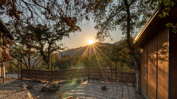 Een terras met houten stoelen, een vuurkuil en uitzicht op de ondergaande zon achter heuvels.
