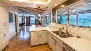 Een moderne keuken met houten vloeren, witte kasten, een groot eiland met een spoelbak en een uitzicht op de omgeving door grote ramen.
