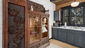A kitchen with dark gray cabinets, a wooden cabinet with intricate carvings, a microwave, and a large window with a view of trees. - Agave | Fireplace | Outdoor Shower (Fredericksburg)