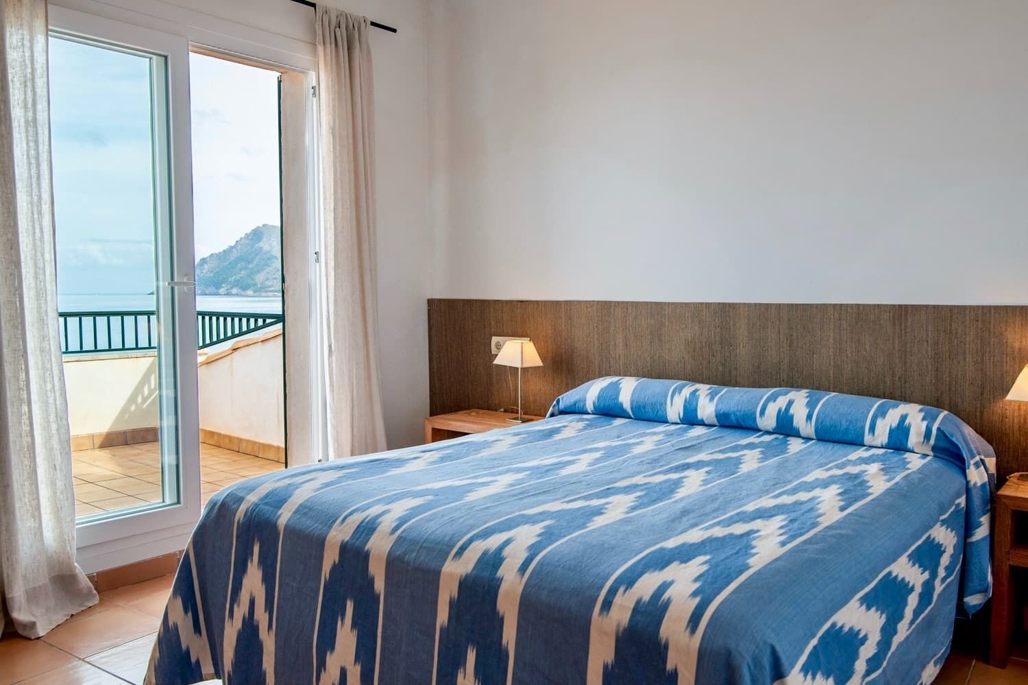Un dormitorio con cama, una mesita de noche, una lámpara y vista al mar a través de una puerta corrediza de vidrio.