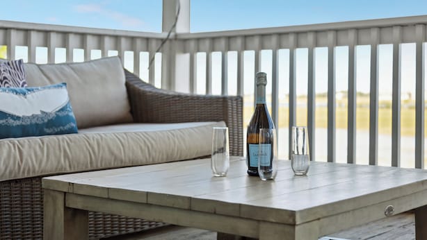 Ein Korbsessel mit Kissen, ein Holztisch und zwei Gläser auf einem Balkon mit Geländer.