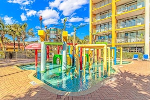Un espacio de juegos acuáticos con estructuras coloridas, una piscina y un edificio de varios pisos con balcones.