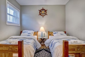 Una habitación con dos camas de madera, un adorno de timón de barco, una lámpara y una ventana con persianas.
