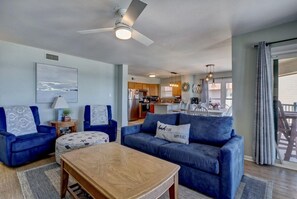 A living room with a sofa, ottoman, and coffee table, a ceiling fan, and a view into the kitchen area. - Top Floor Oceanfront & Sound Views, Pool, (North Topsail Beach)