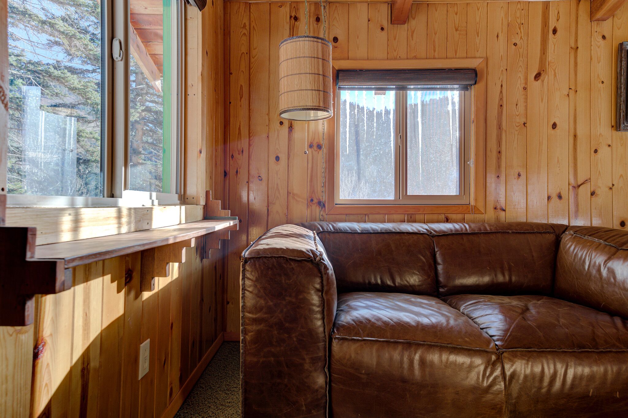 Interno di una baita in legno con un divano in pelle marrone, una finestra con vista sugli alberi e un lampadario.