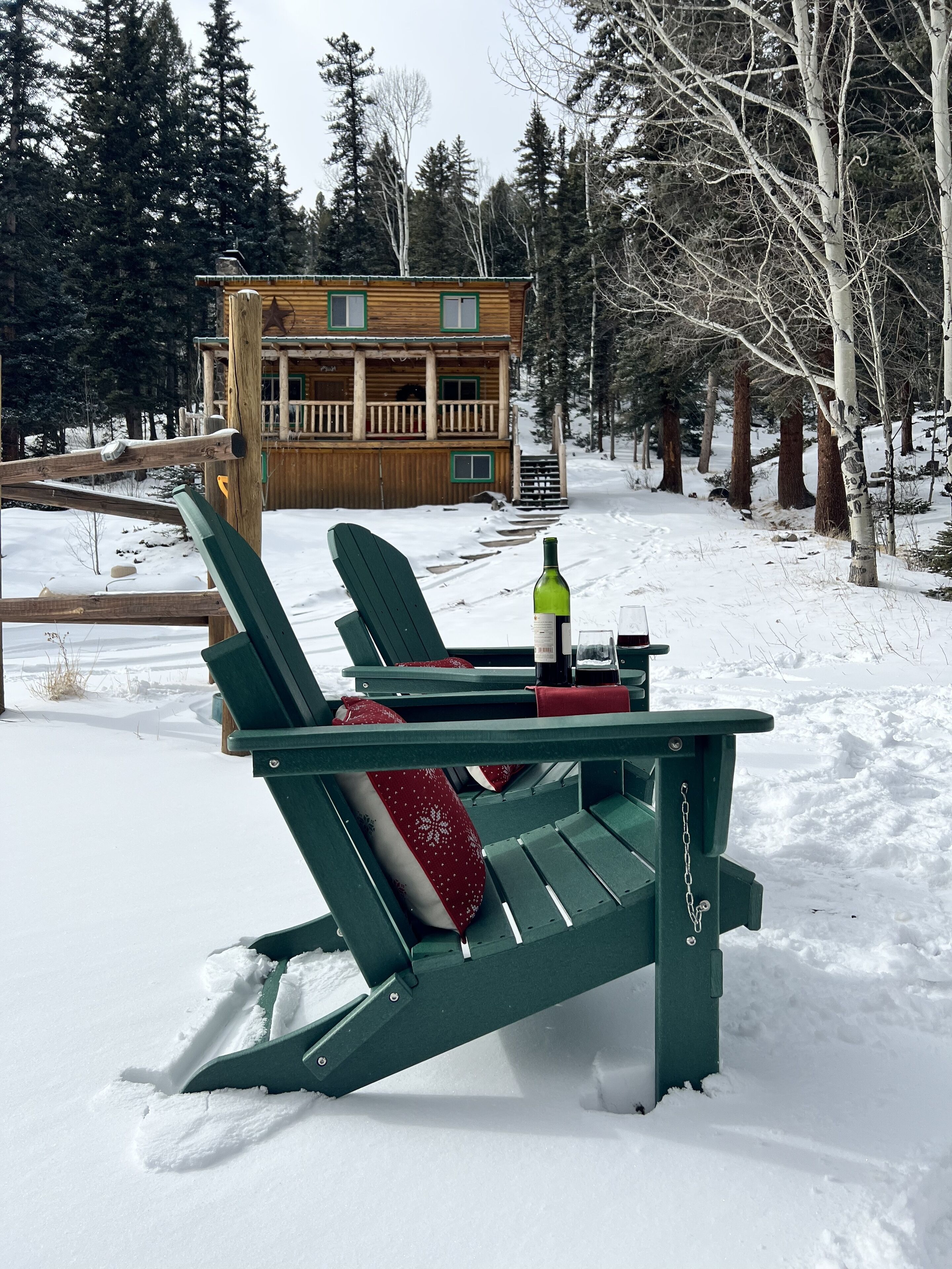 A snow-covered outdoor area with a wooden cabin in the background, a green Adirondack chair with a red cushion, and a bottle and glass on the chair.