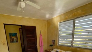 A bedroom with a bed, a ceiling fan, a window with blinds, a door, and a small table with a lamp. - Best Location, Caribbean views, Off street parking, walk everywhere. (Vieques)
